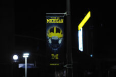 A Michigan football banner is illuminated by street lights with the stadium's giant scoreboard in the background.
